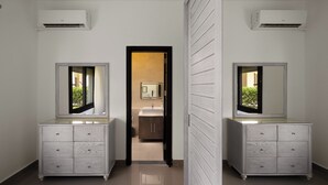 A mirrored room with two white dressers, a bathroom with a sink and mirror, and an air conditioning unit on the ceiling.