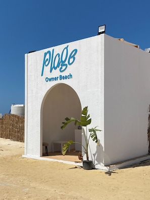 Un bâtiment sur lequel on peut lire l’inscription « Plage Owner Beach », avec une façade blanche, une entrée en arc de cercle et un pot de fleurs.