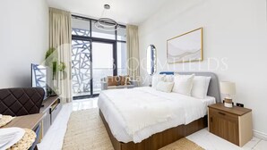 A modern hotel room with a large bed, a bedside table with a lamp, a dining area with a table and chairs, and a view of the city through large windows. - Bloomfields Homey Stu The Gate Masdar  (Abu Dhabi)