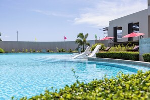 Piscina con sillas de descanso y sombrillas, rodeada de vegetación y un edificio moderno.