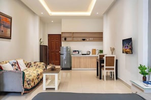 Ein modernes Wohnzimmer mit einer Couch, einem Couchtisch und einem Essbereich mit einem Stuhl und Tisch. Im Hintergrund ist eine Küche mit Kühlschrank und Mikrowelle zu sehen.