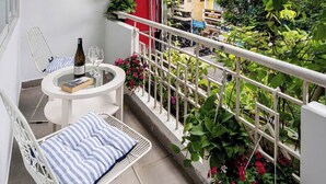 Un balcón con una silla blanca, una mesita pequeña, una botella y un vaso. Hay flores rojas y plantas verdes.