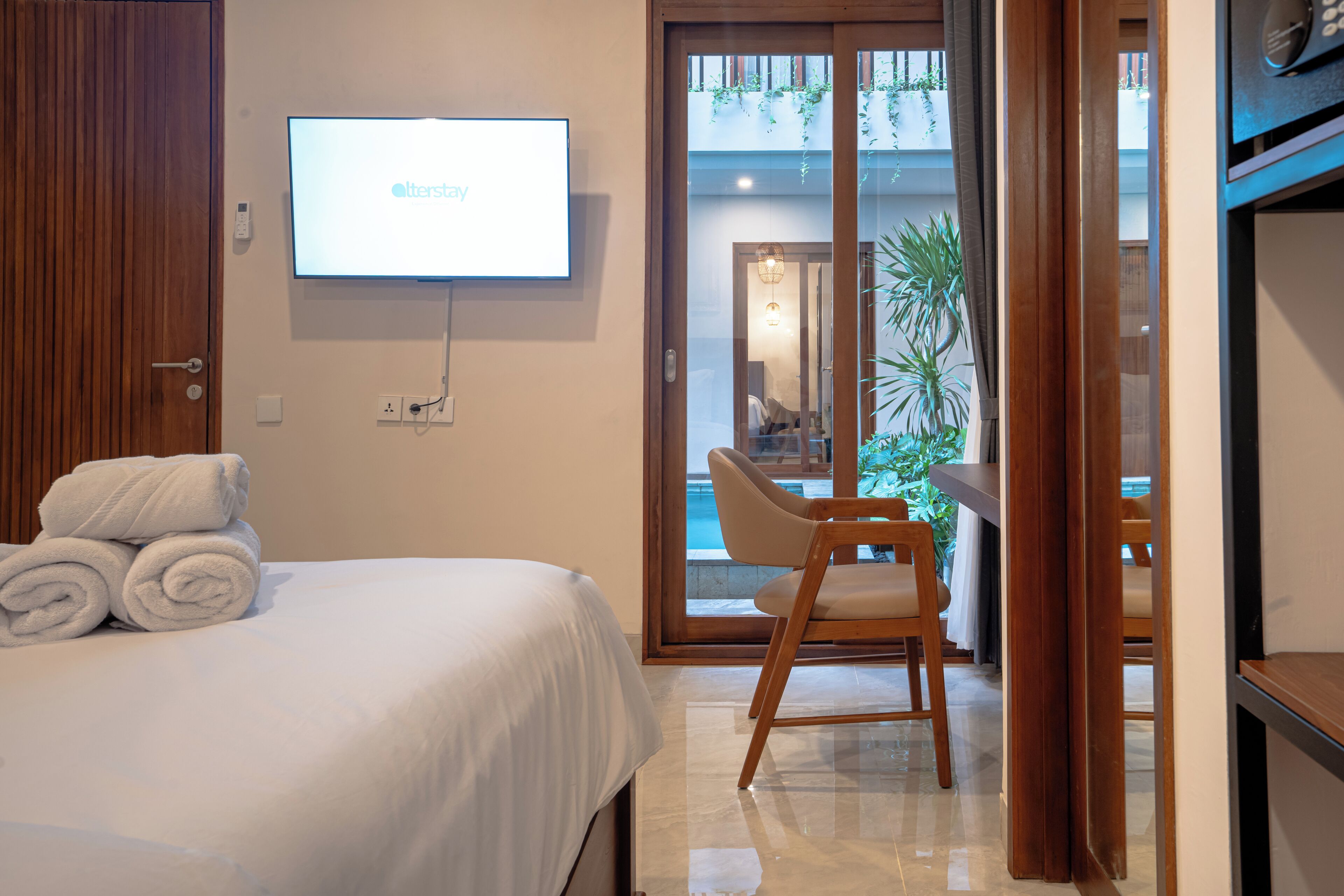 A hotel room with a bed, a TV mounted on the wall, a chair, and a view of the pool through a glass door.