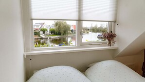 A bedroom with a window overlooking a canal, a bed with floral bedding, and a small vase with dried flowers on the windowsill. - Nice holiday home on the water in Grou (Grou)