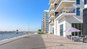 A waterfront promenade with modern buildings, outdoor seating, and a clear view of the water. - FIRST CLASS | 1BR | Tranquility in Style (Abu Dhabi)