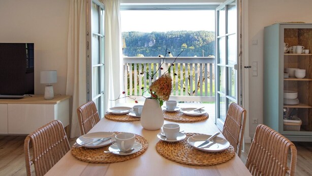 A dining area with a table set for four, a television on a stand, and a view of a forested landscape through an open door. - Fantastic guest house on the Elbe cycle path and Bastei (Rathen)