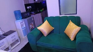 Un canapé vert agrémenté de deux coussins dorés, une petite table blanche avec des tiroirs, un distributeur d’eau et un téléviseur posé sur un meuble.