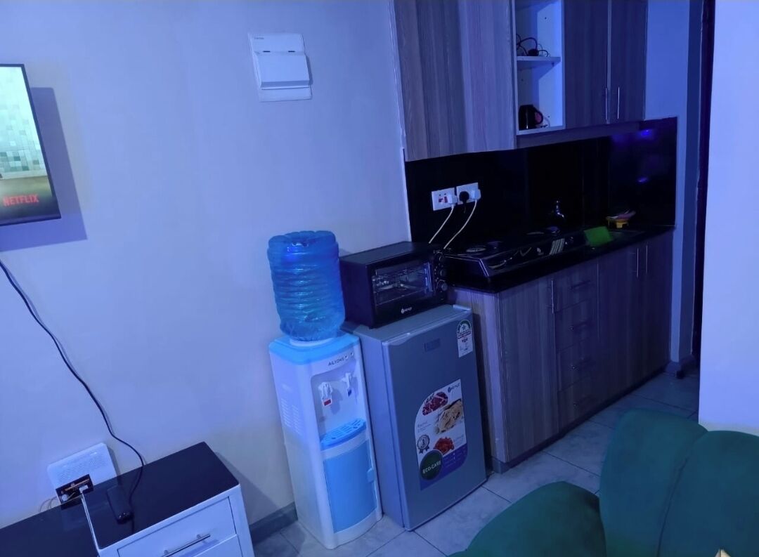 A compact living space with a kitchenette, a water dispenser, a small refrigerator, and a TV mounted on the wall.