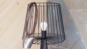 En moderne lampe med metalramme og hvid pære, stående på en træoverflade med en stenvæg i baggrunden.