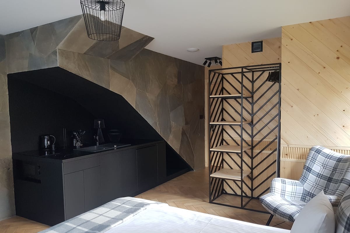 A modern bedroom with a bed, a black kitchenette, a wooden wardrobe, and a plaid chair.