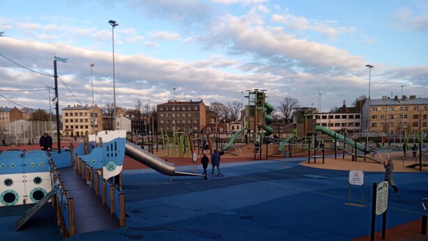 An outdoor playground with slides and climbing structures, surrounded by buildings.