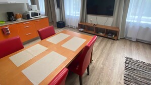 A modern living room with a wooden dining table, red chairs, a flat-screen TV mounted on the wall, and a kitchen area with a microwave and coffee maker.