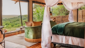 Una habitación con cama, una silla verde, una mesita y un ventanal grande con vistas a un paisaje exuberante.