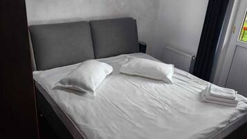 A single bed with white linens and two pillows, a grey headboard, a radiator, and a window with dark curtains.