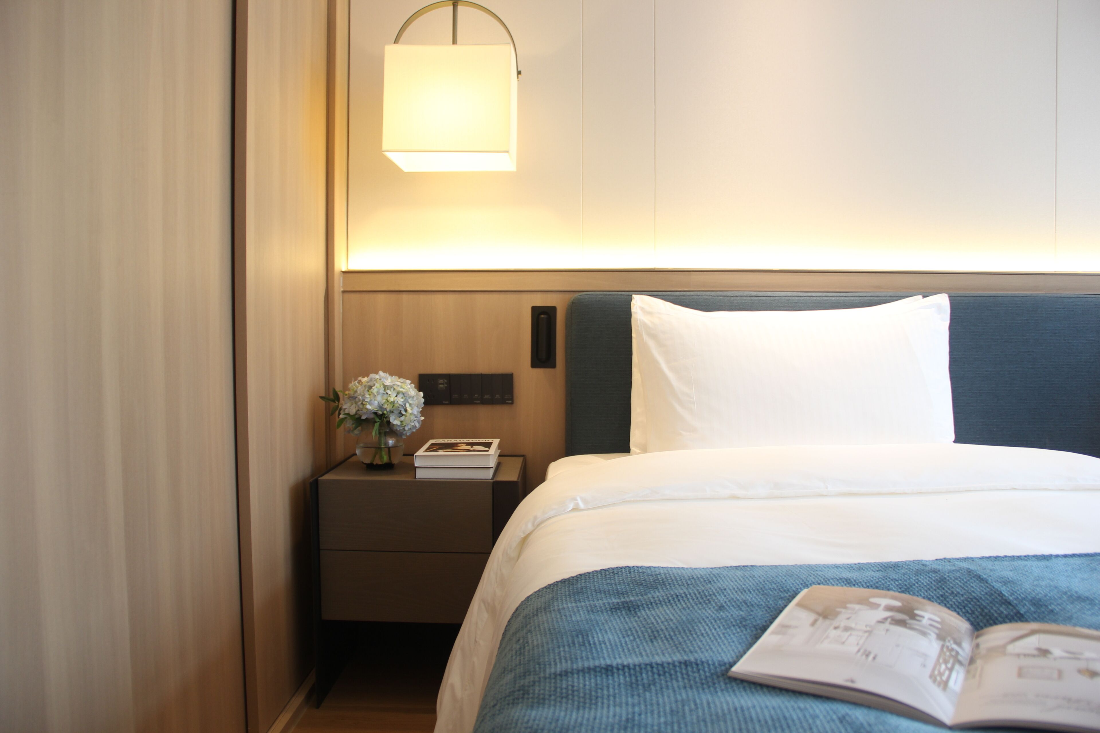 A hotel room with a neatly made bed, a bedside table with a lamp, and a book on the bed.