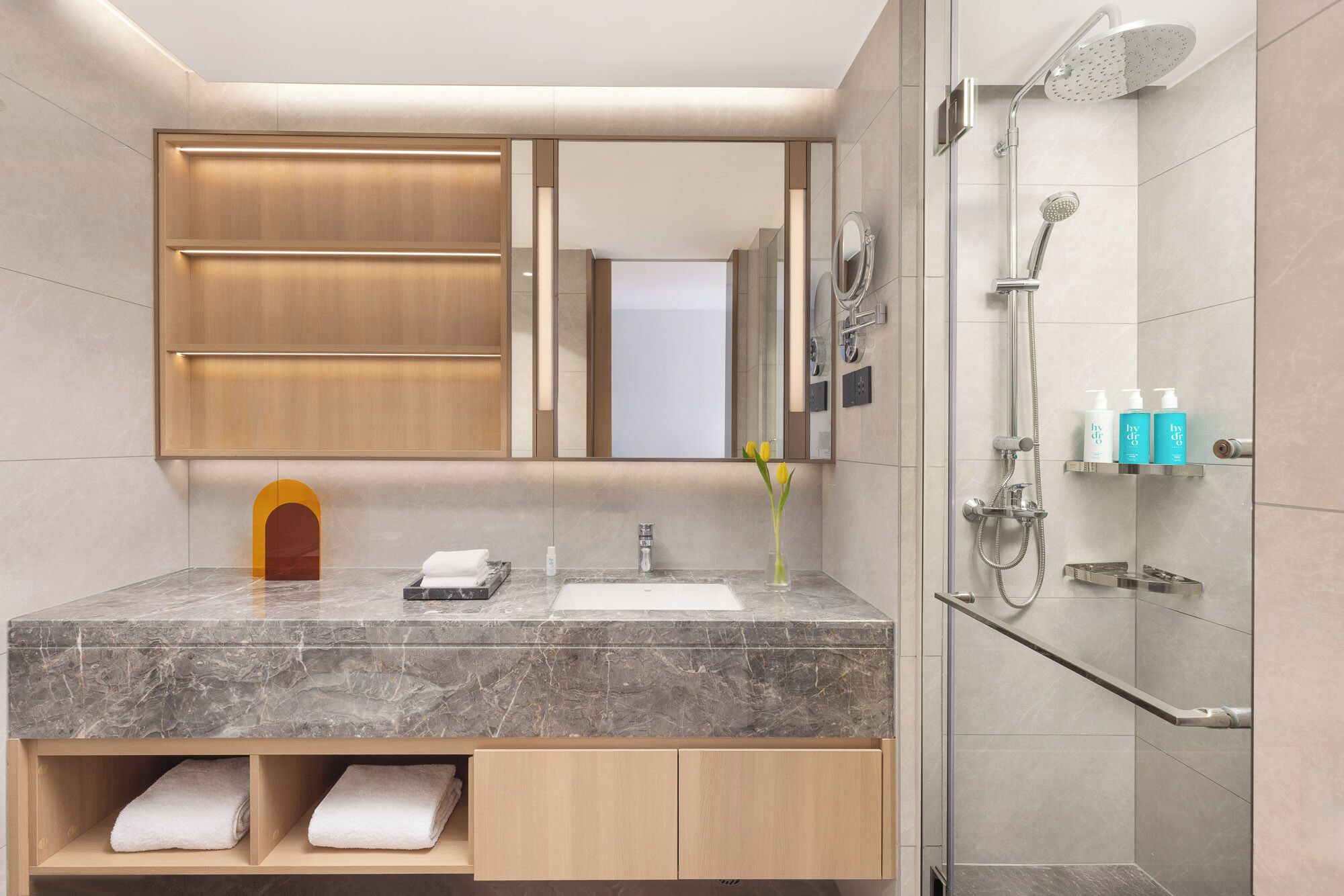 A modern bathroom with a marble countertop, a wooden vanity, a large mirror, a shower with a handheld showerhead, and a shelf with toiletries.