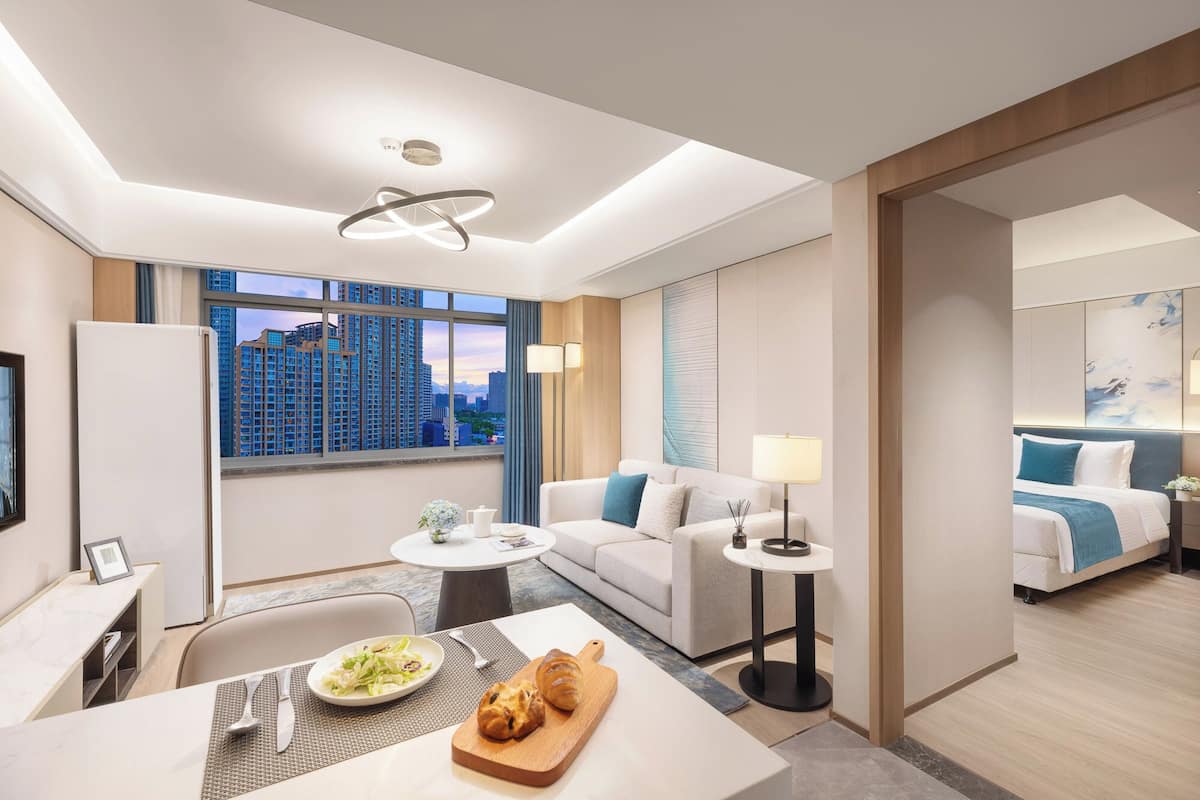 Une chambre d’hôtel moderne avec vue sur la ville, comprenant un coin repas, un canapé, un lit et une petite table sur laquelle se trouve une assiette de nourriture.