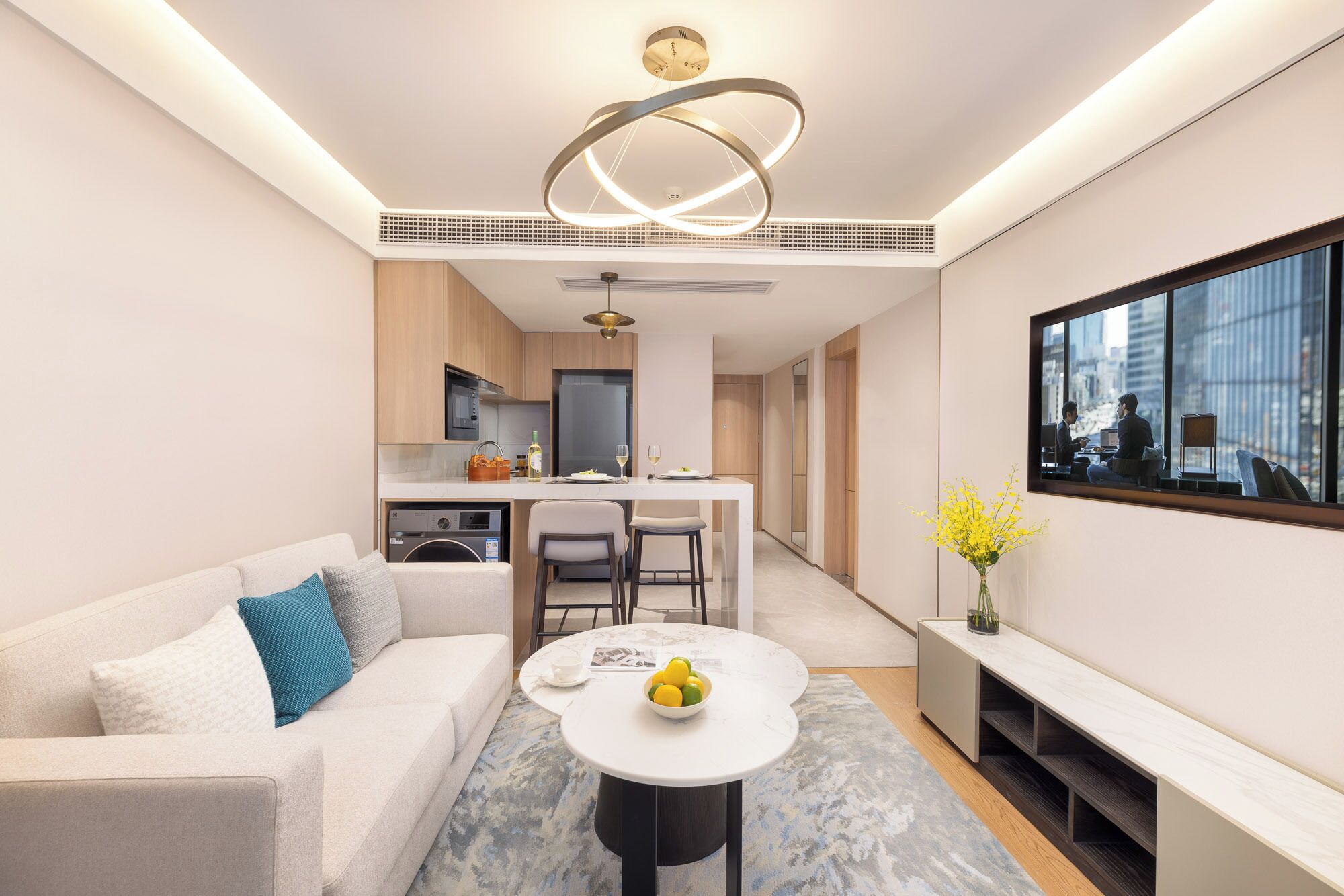 A modern living room with a sofa, a coffee table with fruit, a TV, and a kitchen in the background.