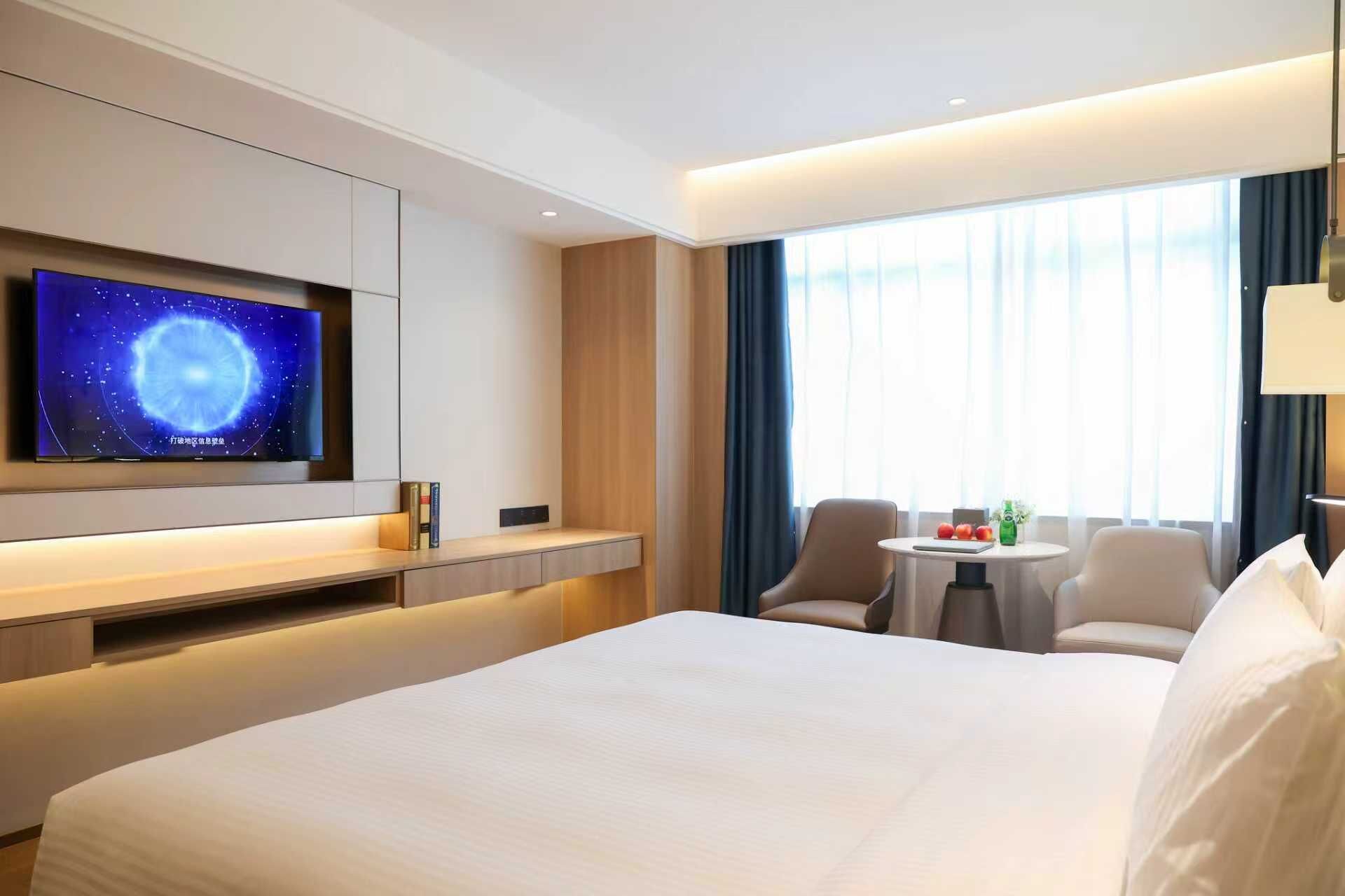 A hotel room with a large bed, a TV mounted on the wall, a small table with chairs, and a window with curtains.