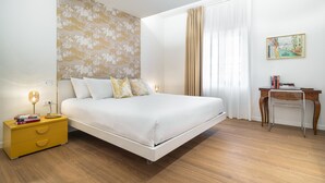 A modern bedroom with a bed, a bedside table with books, a desk, and a painting on the wall.
