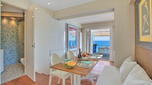 Una habitación de hotel moderna con una mesa de comedor de madera, sillas blancas, un sofá blanco y un balcón con vistas al mar.
