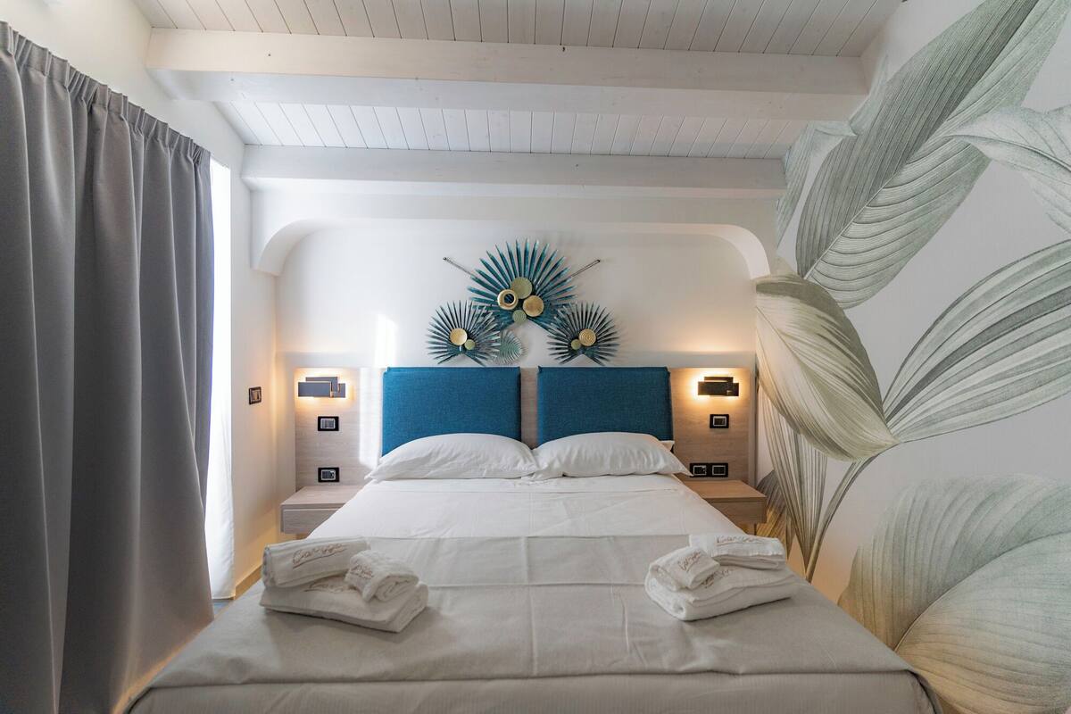 A hotel room with a large bed, a headboard with a decorative sunburst design, bedside lamps, and a large leaf-patterned wall.