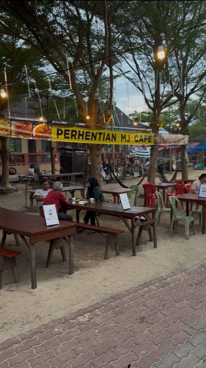 An outdoor cafe with wooden tables and chairs, a yellow sign reading "PERHENTIAN MJ CAFE", and trees providing shade.