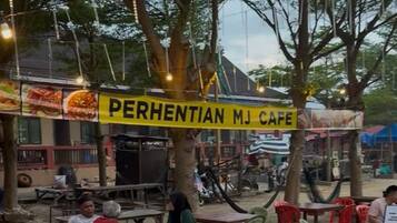 An outdoor cafe with wooden tables and chairs, a yellow sign reading "PERHENTIAN MJ CAFE", and trees providing shade.