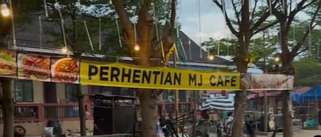 Un café al aire libre con mesas y sillas de madera, un letrero amarillo que dice "PERHENTIAN MJ CAFE" y árboles que dan sombra.