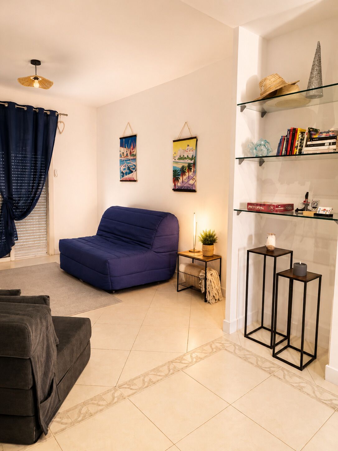 Een woonkamer met een blauwe bank, een klein bijzettafeltje met een plant, een boekenkast met boeken en decoratieve objecten, en twee aan de muur bevestigde afbeeldingen.