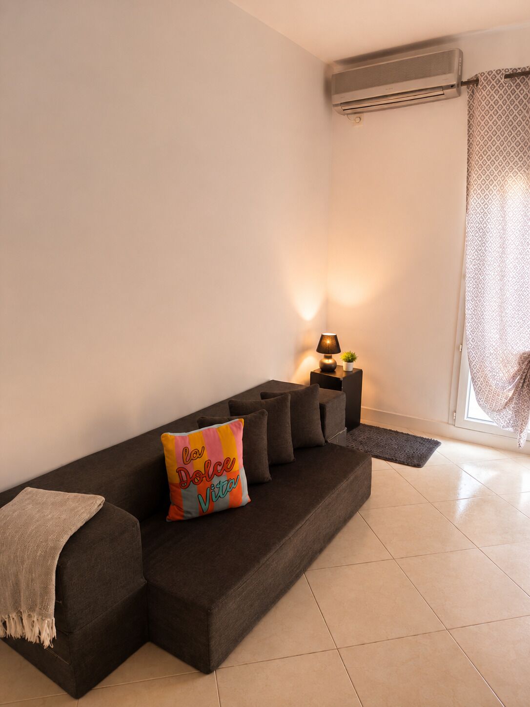 A modern living room with a dark sofa, a decorative pillow, a small table with a lamp, and an air conditioning unit on the wall.