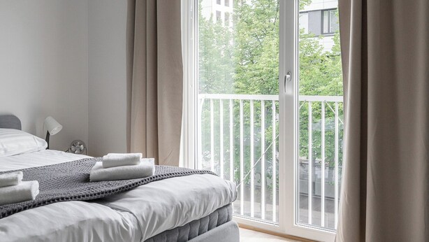 Un dormitorio moderno con cama, mesita de noche, lámpara y vistas a un balcón con cortinas.