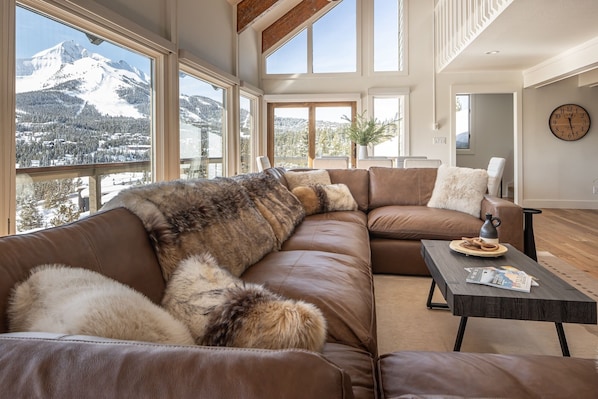 A spacious living room with a large brown leather sectional sofa, a coffee table, and a view of snow-covered mountains through large windows. - Game Room | Hot Tub | Near Skiing (Big Sky)