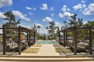 A covered outdoor seating area with lounge chairs and umbrellas, a pool, and palm trees in the background.
