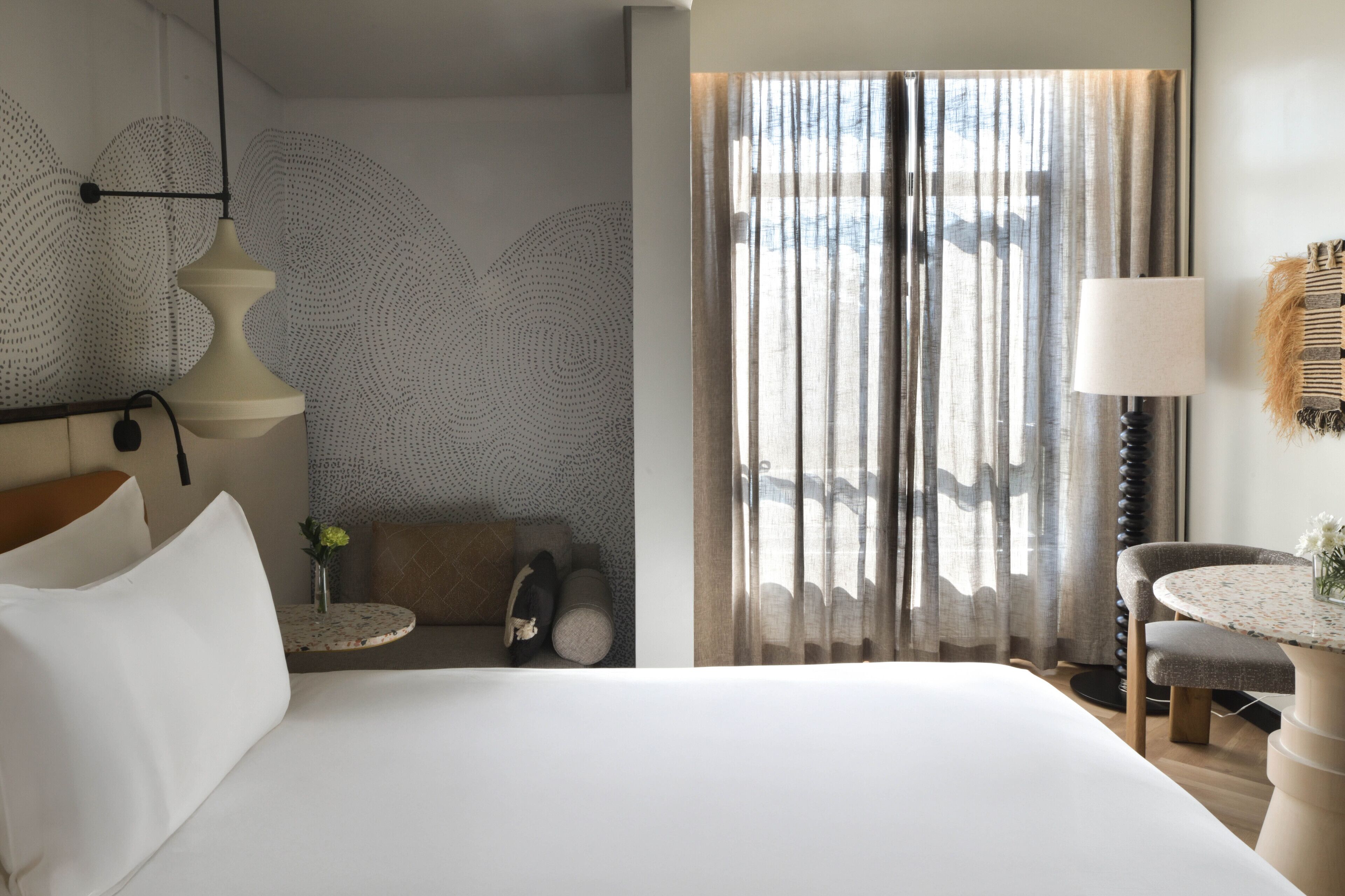 Une chambre d’hôtel avec un grand lit, une table ronde, une chaise, une lampe et une vue sur l’extérieur à travers des rideaux légers.