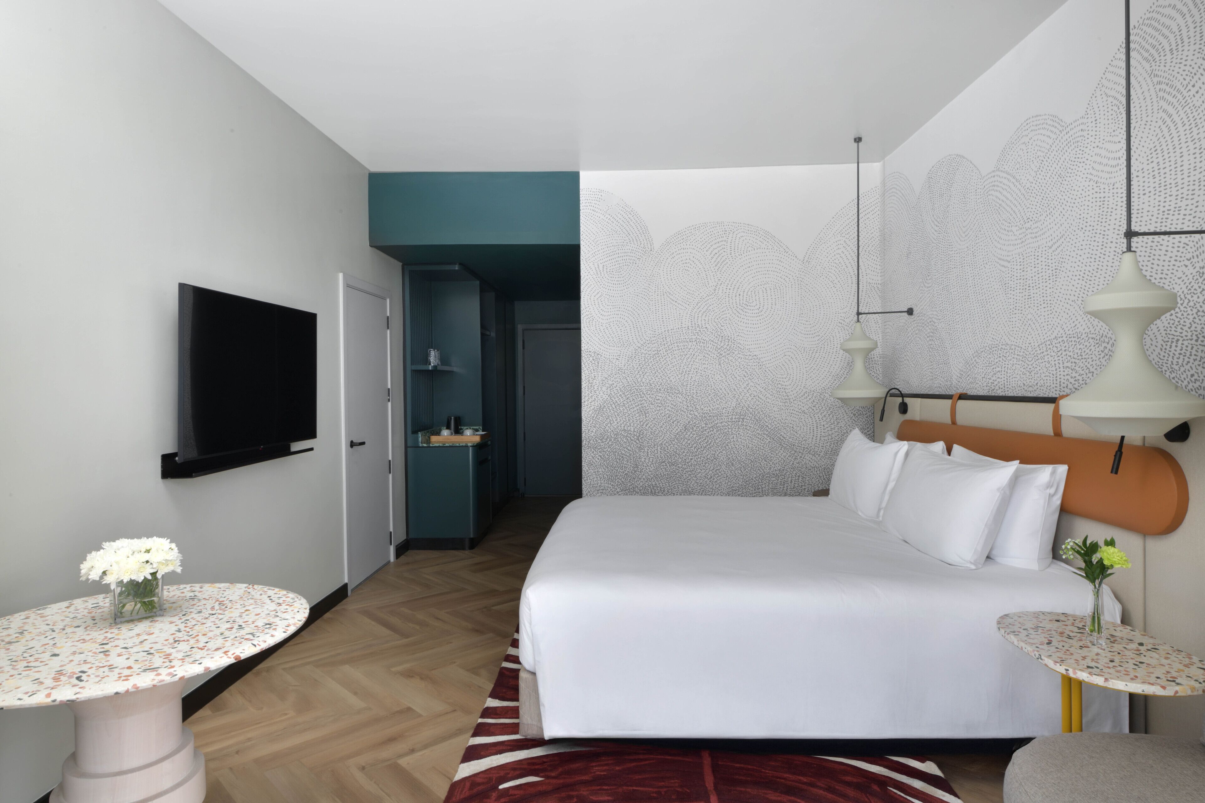 Quarto de hotel moderno com uma cama grande, uma televisão fixada na parede, uma mesa pequena com um vaso de flores e um papel de parede estampado.