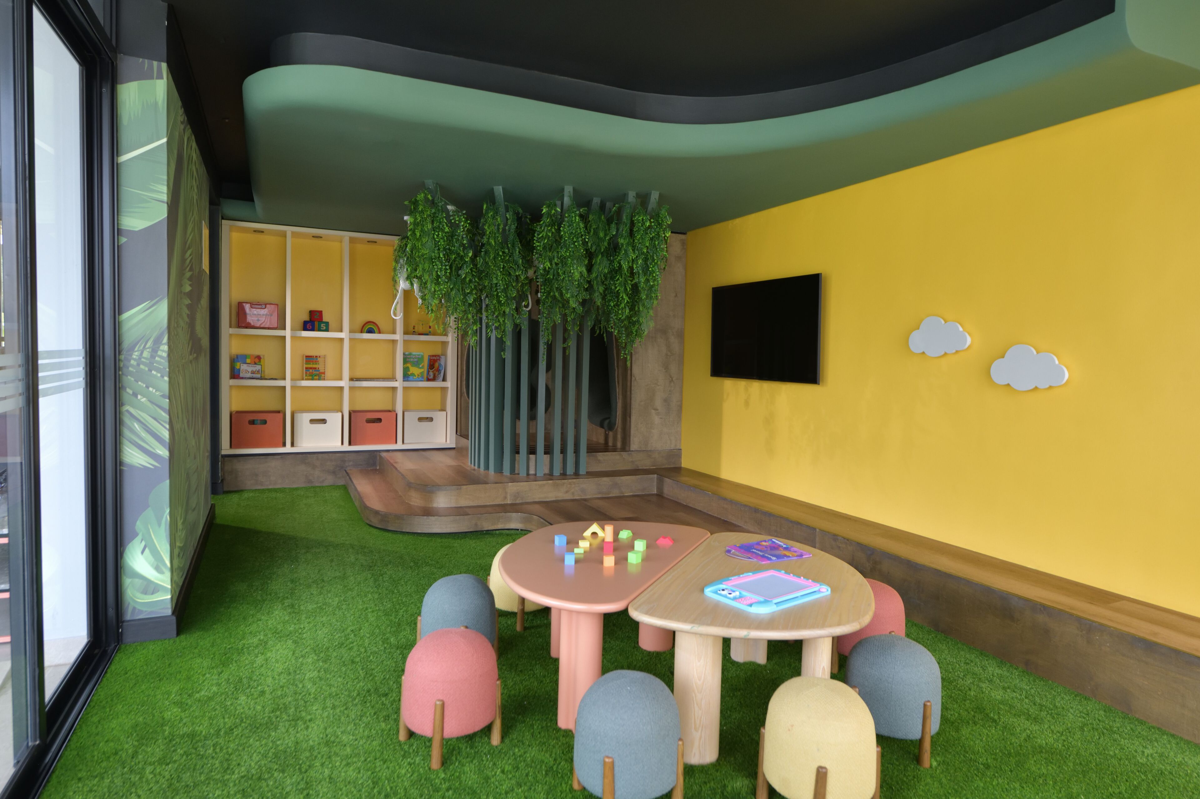 Una habitación con césped artificial, una zona de juegos con juguetes, una estantería y un televisor de pared.