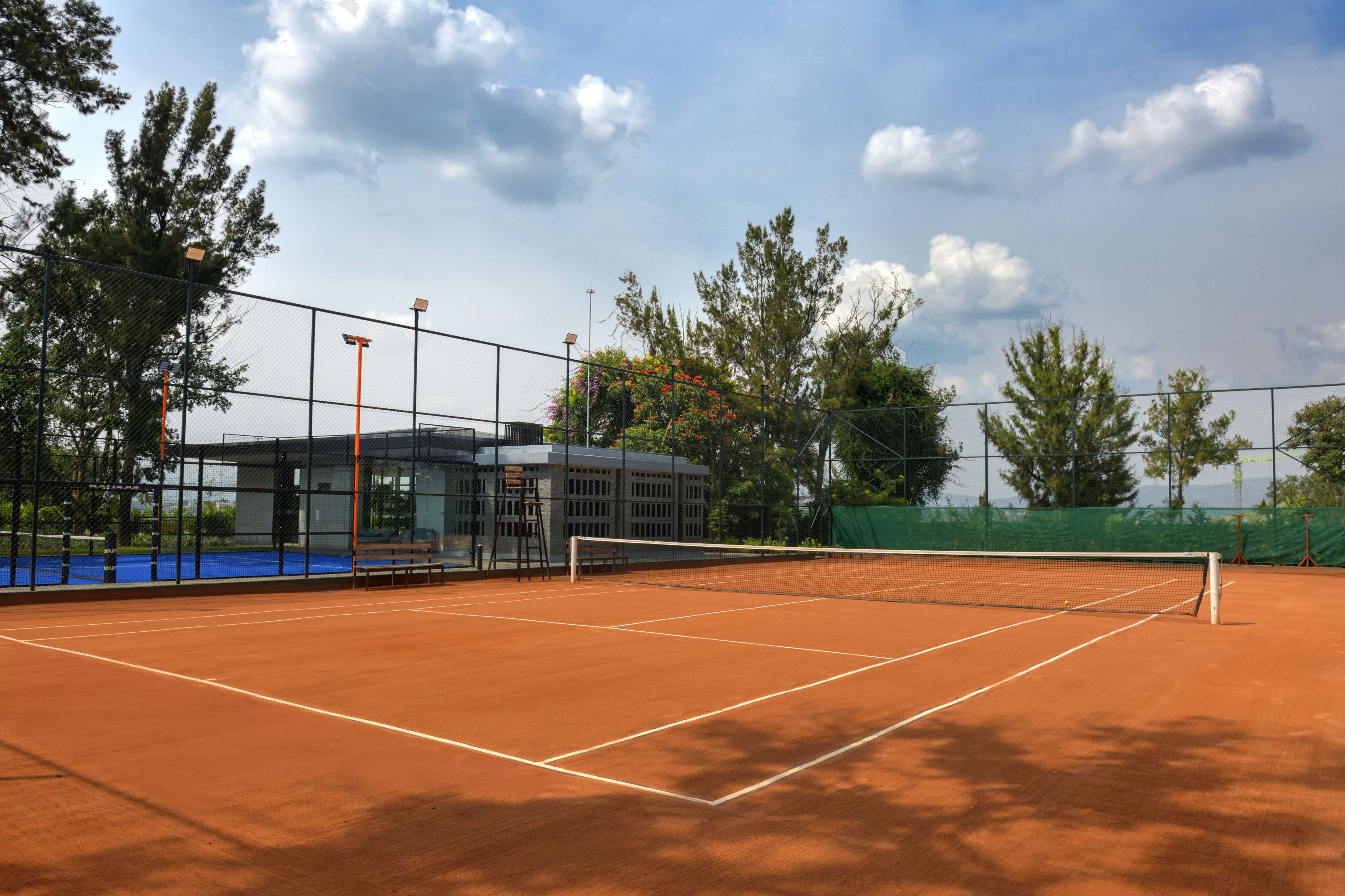 Una cancha de tenis con red, rodeada por una cerca, y un edificio con ventanas grandes al fondo.
