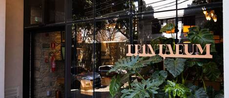 Una entrada con fachada de vidrio y un letrero que dice 'TRINTEUM', con plantas verdes grandes en el frente.