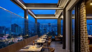 Un restaurant situé sur le toit d’un immeuble, avec des tables et des chaises, une vue sur la ville et un rideau à motifs.