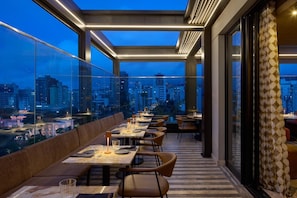 Un restaurante en la azotea con mesas y sillas, vistas a la ciudad y una cortina estampada.