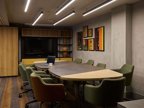 Un salón de conferencias con una gran mesa, sillas verdes, una computadora portátil y un televisor en un estante.