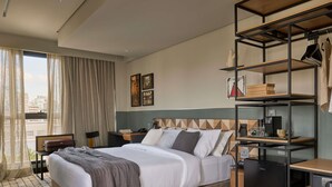 Una habitación de hotel moderna con una cama grande, un escritorio con cafetera, una estantería y vistas a la ciudad desde la ventana.
