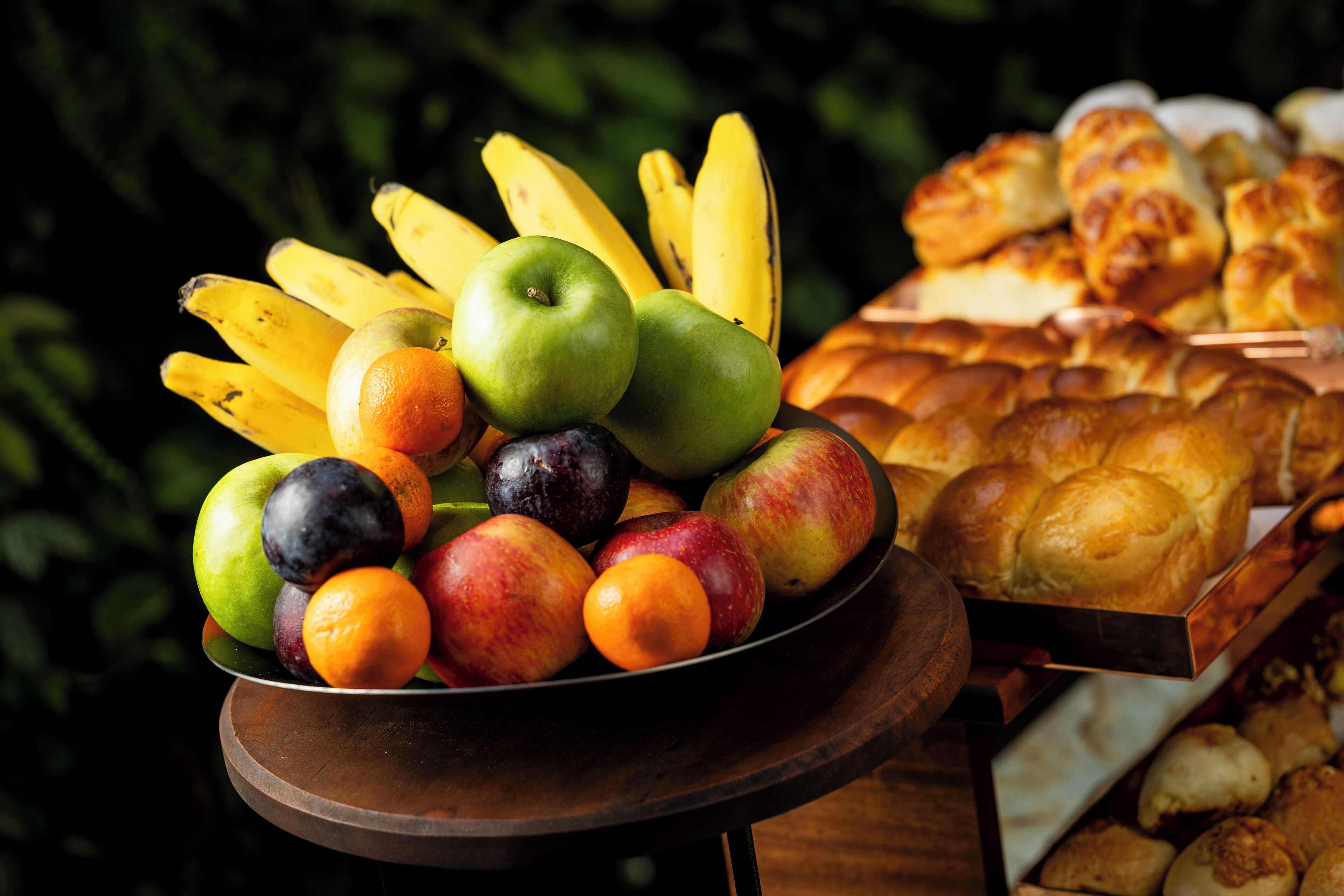 Un frutero con manzanas, naranjas y plátanos, junto a una bandeja de pan.