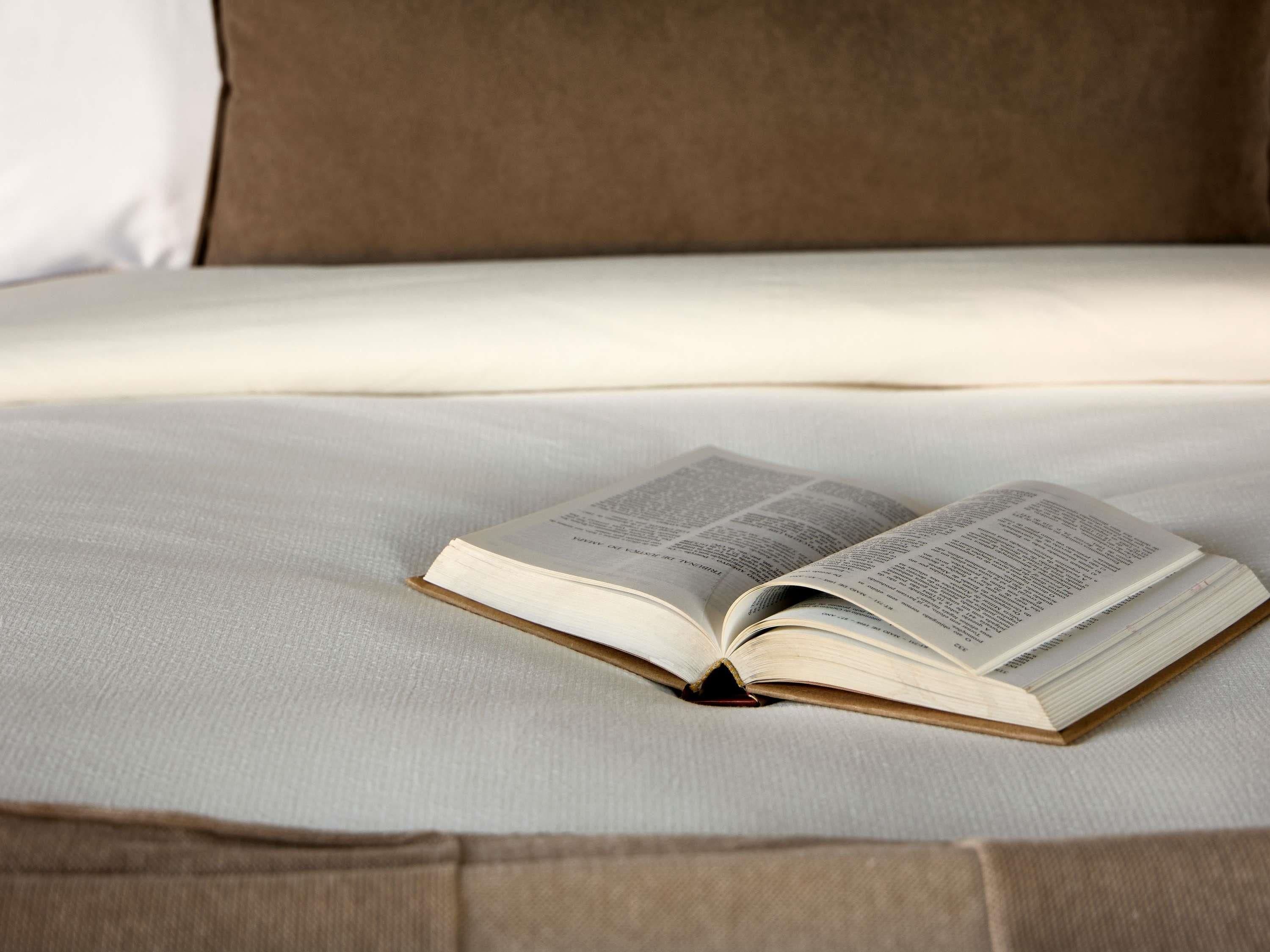 An open book on a bed with a plain white sheet and a brown pillow.