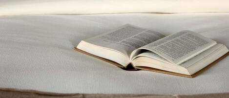 An open book on a bed with a plain white sheet and a brown pillow.