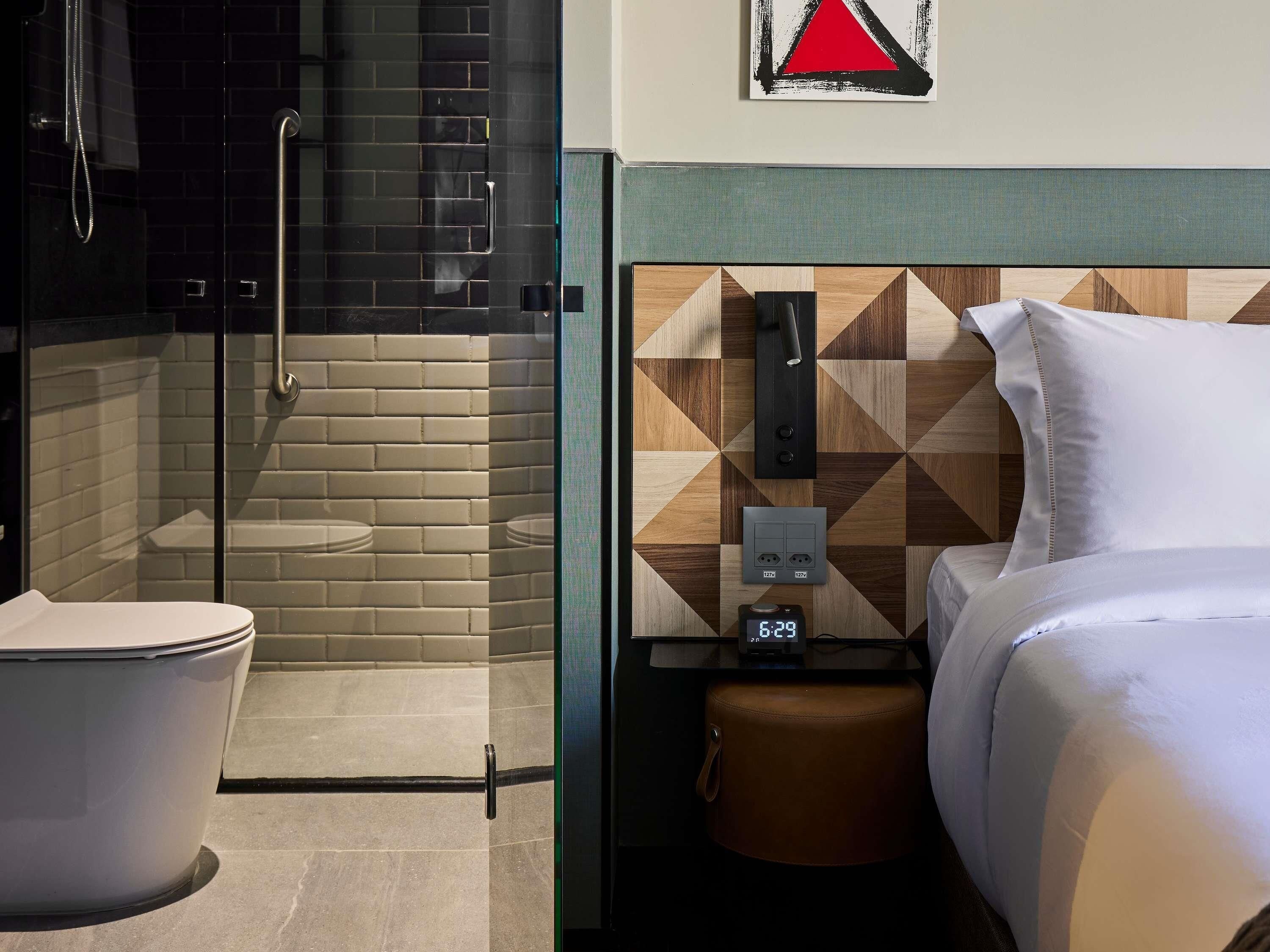 Habitación de hotel con ducha, inodoro, cama, mesita de noche y un reloj de pared.