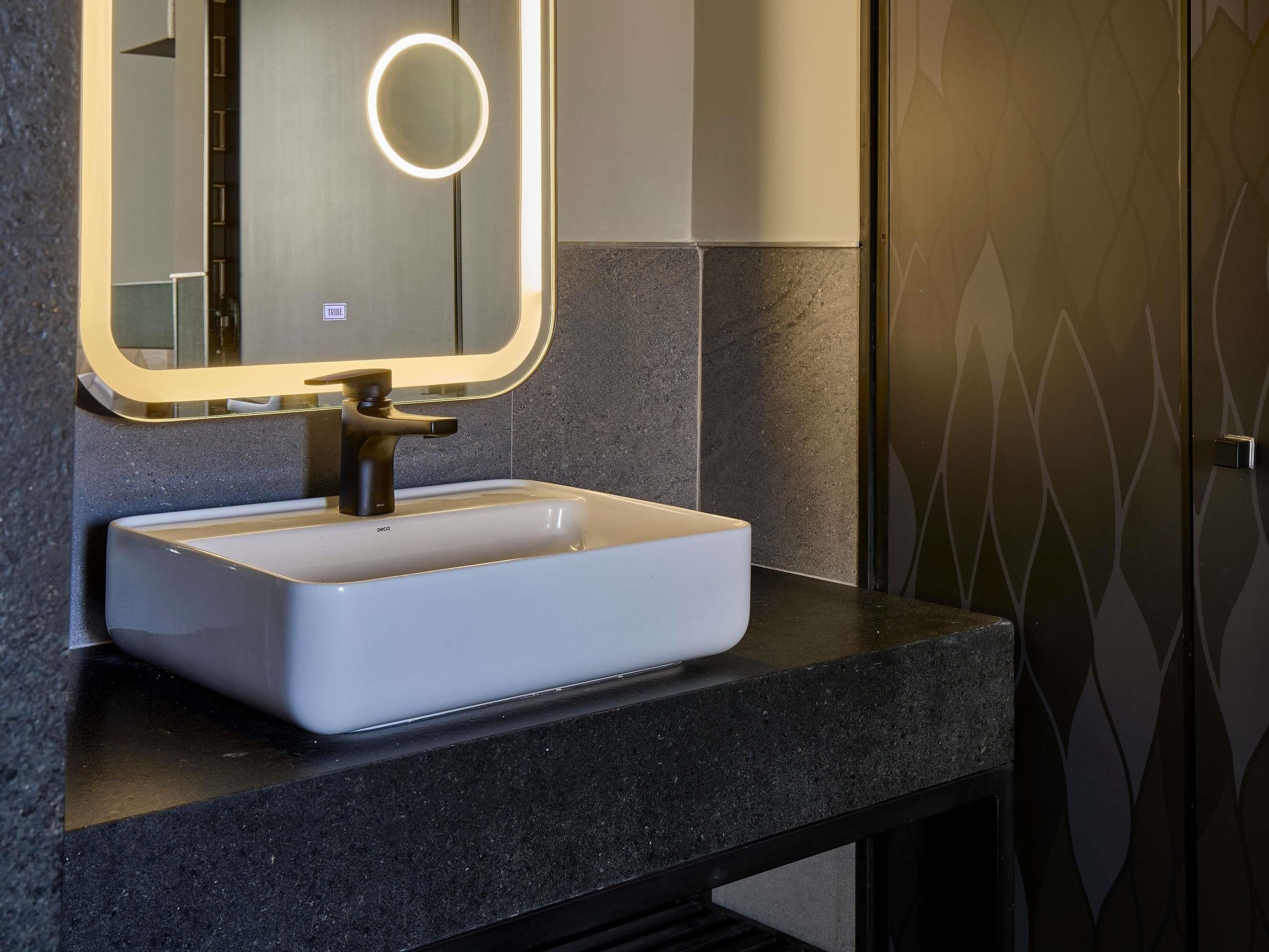 Un baño con un lavabo blanco, un espejo con luz circular y una pared de azulejos oscuros.