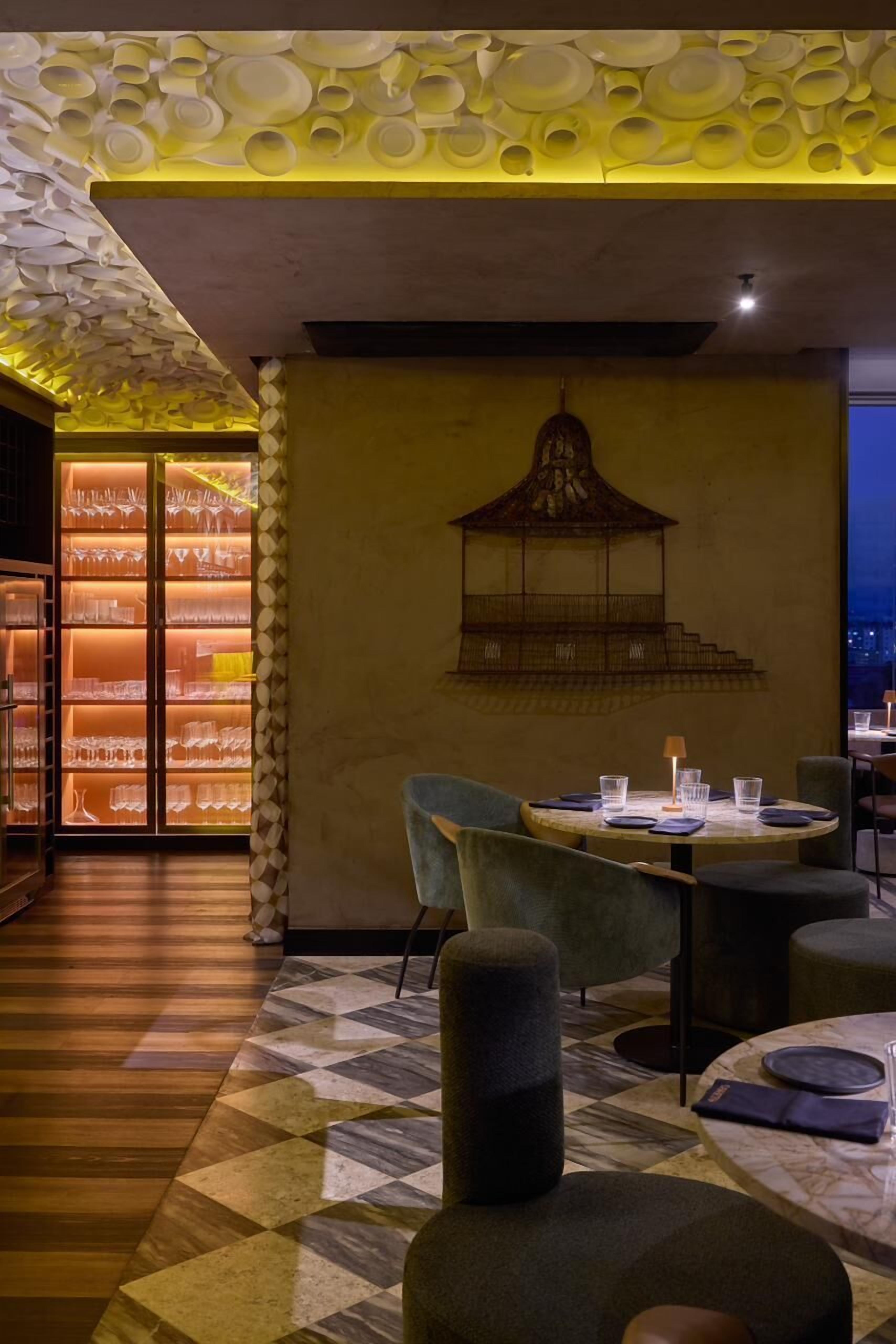Interior de restaurante com teto estampado, obra de arte com gaiola de pássaro na parede e área de refeições com mesas e cadeiras redondas.