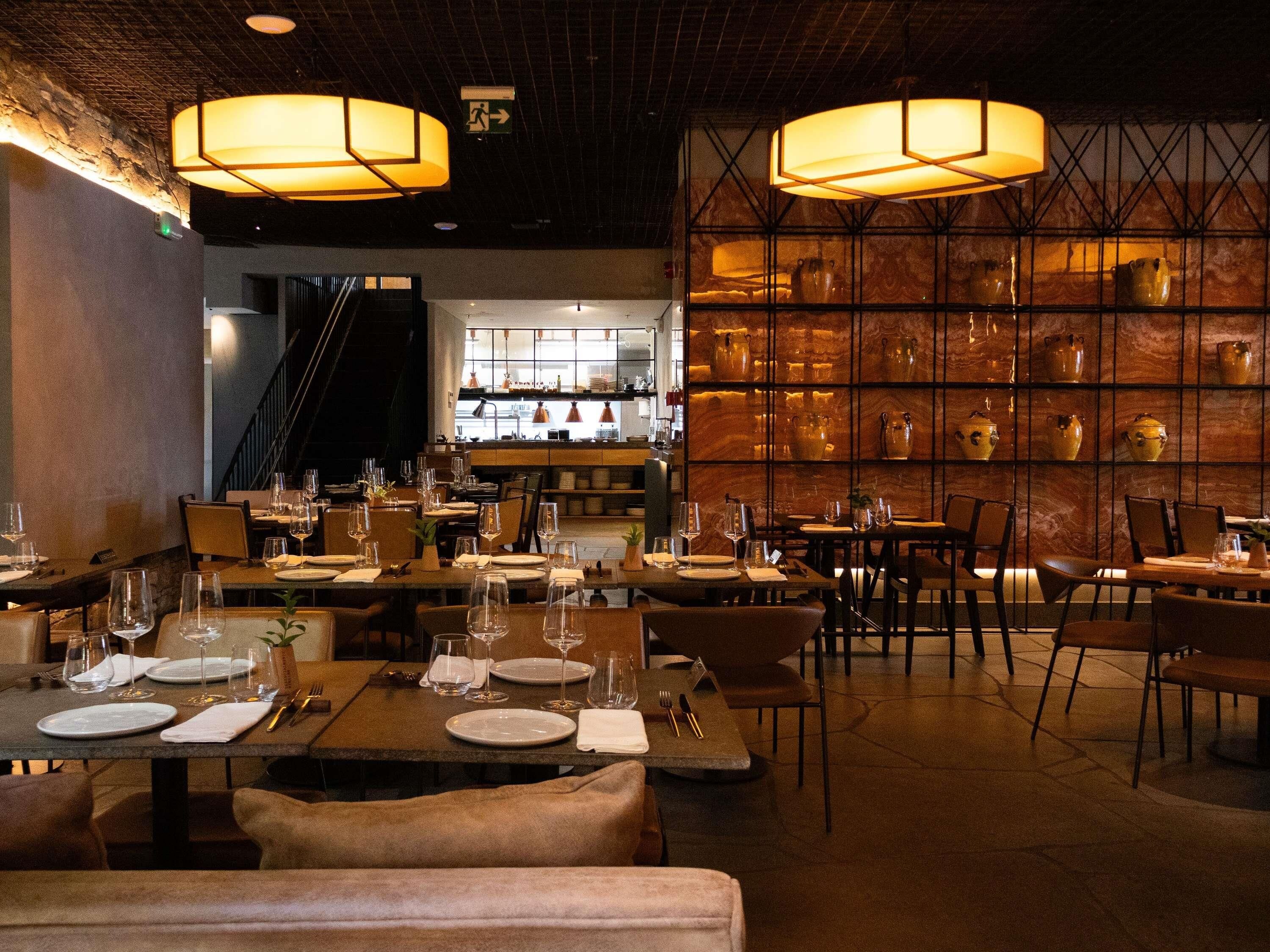 A restaurant interior with tables set for dining, a bar area in the background, and decorative wall shelves.